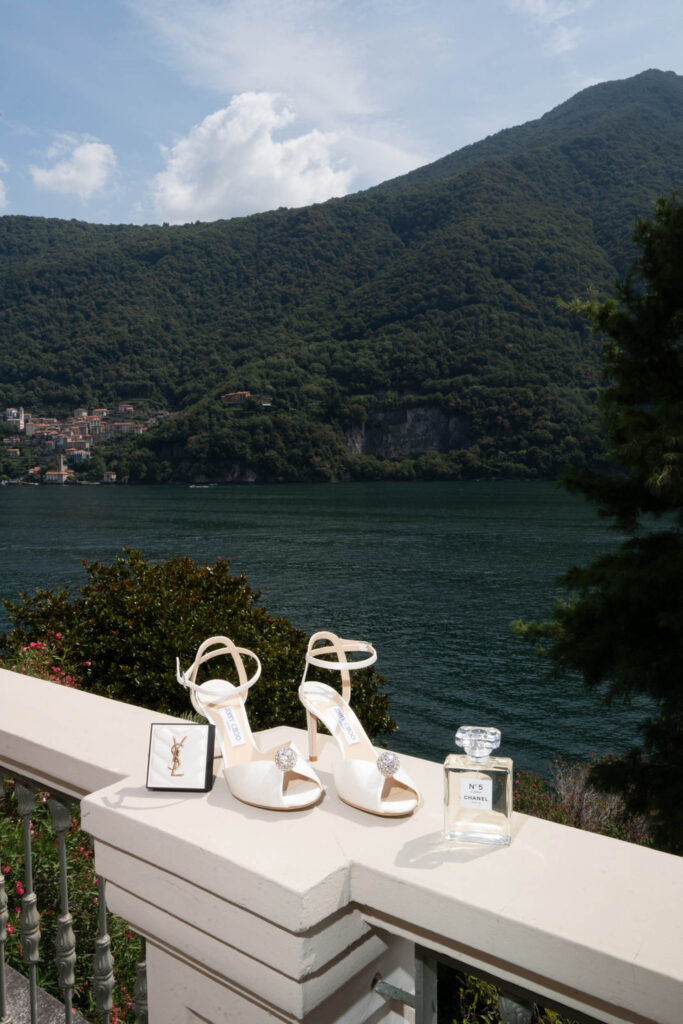 Weiße High Heels, eine YSL-Schachtel und Parfüm, elegant arrangiert auf einem Balkon mit Blick auf den atemberaubenden Comer See und die Berge in Italien.