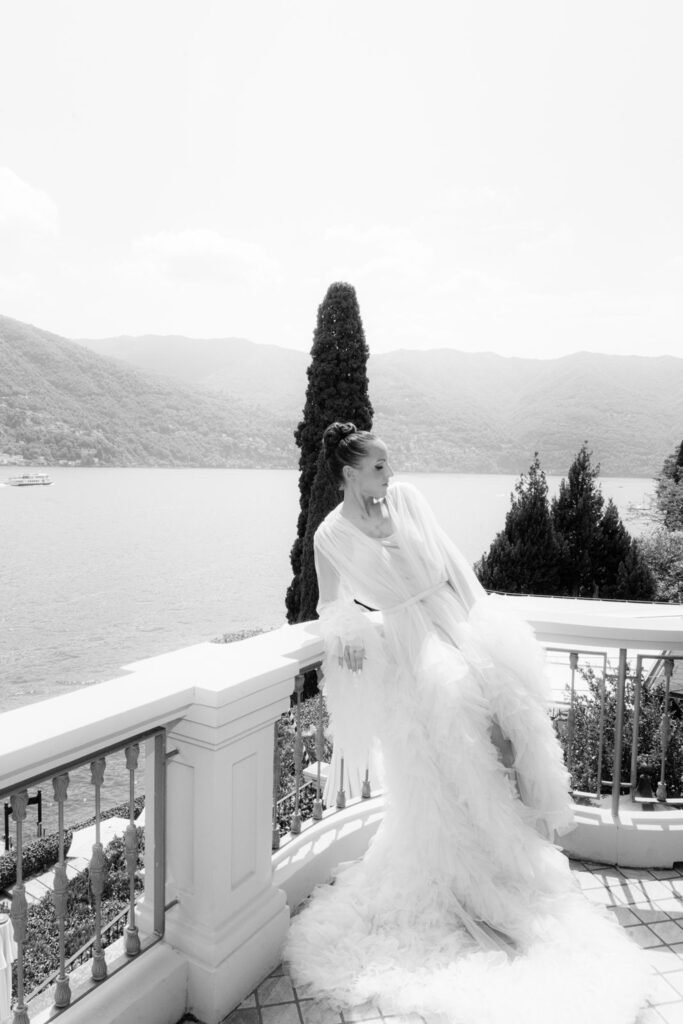 A woman in a flowing white dress stands on a balcony overlooking Lake Como and the mountains; the black-and-white image conveys the timeless elegance of the Italian landscape.