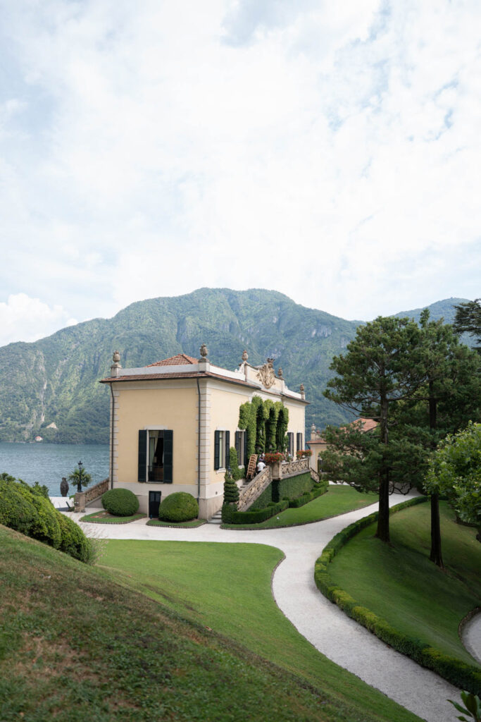 Eine gelbe Villa mit Efeu, direkt am See, vor einer Bergkulisse und mit einem gewundenen Weg durch gepflegte grüne Rasenflächen und Bäume – wie ein automatisch gespeicherter Entwurf eines ruhigen Rückzugsortes inmitten der Natur.
