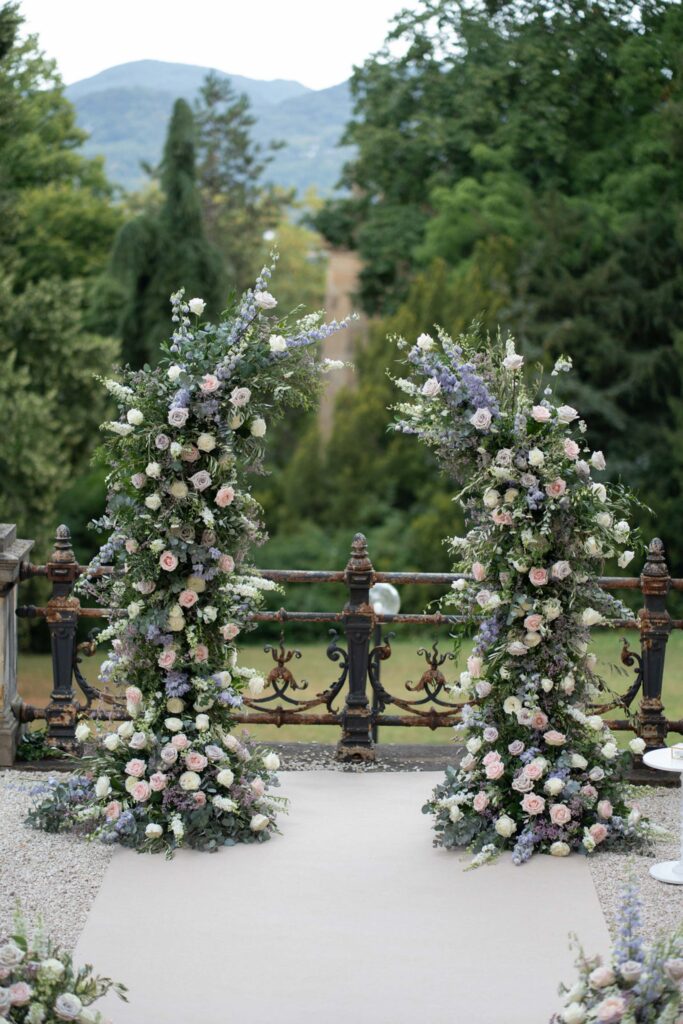 Zwei hohe Blumengestecke aus weißen und lavendelfarbenen Blüten, im Freien arrangiert, mit Bäumen und Bergen im Hintergrund – perfekt für eine romantische Stella-Rheni-Hochzeit in Bonn oder alle, die in Bonn in einer atemberaubenden Naturkulisse heiraten möchten.