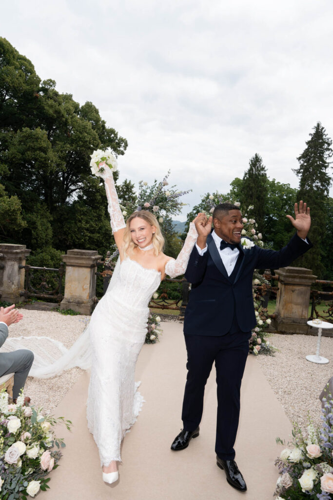Braut und Bräutigam feiern fröhlich im Freien im Stella Rheni, halten Händchen und lächeln, umgeben von Grün und Blumen – eine perfekte Kulisse für Ihre Hochzeit in Bonn.
