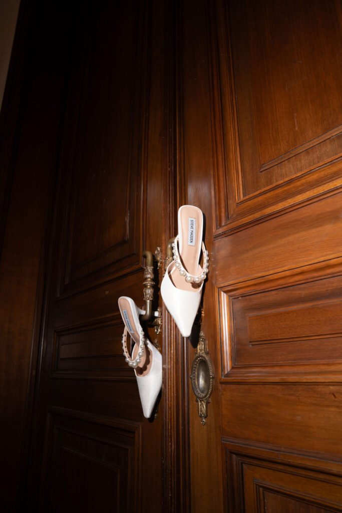 Ein Paar weiße Stöckelschuhe, die an verzierten Messingtürklinken einer Holztür hängen – ein eleganter Moment bei Stella Rheni, perfekt für alle, die von einer Hochzeit in Bonn träumen.