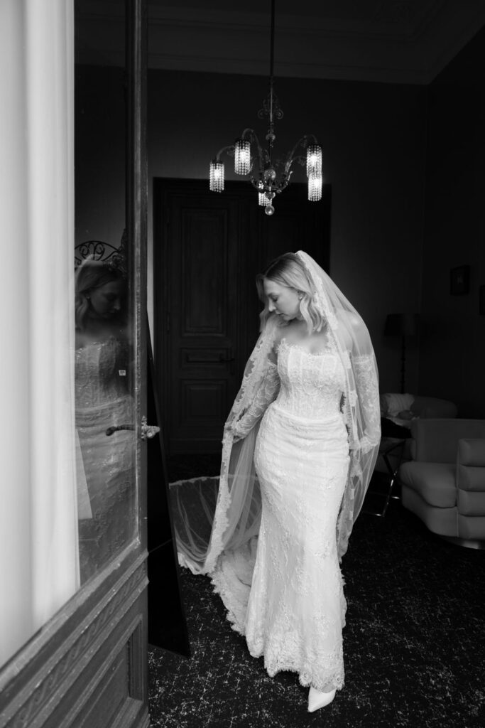 Eine Braut in einem Spitzenkleid und mit Schleier steht in einem Raum, den Blick nach unten gerichtet, die Hand auf die Taille gelegt – ein Bild, das die elegante Atmosphäre einer „Hochzeit Bonn“ einfängt, wie sie sich Stella Rheni vorstellt.