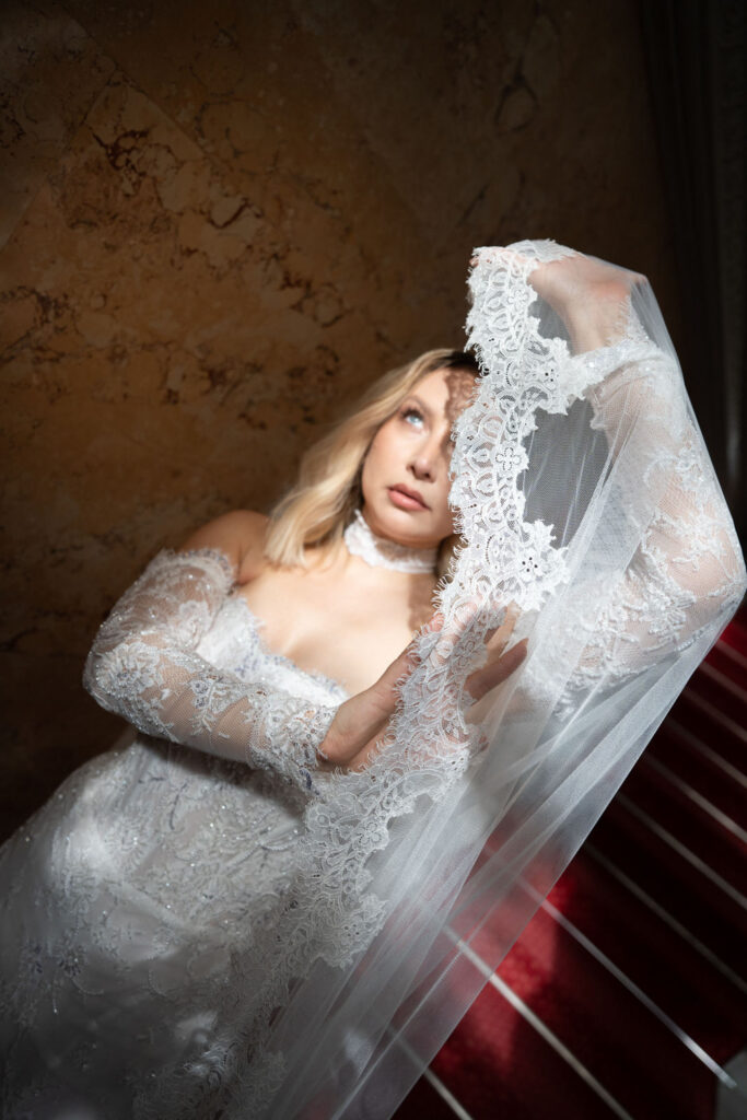 Eine Frau in einem weißen Spitzenhochzeitskleid hält ihren Schleier hoch, während sie auf einer mit rotem Teppich ausgelegten Treppe steht – ein Bild, das die Eleganz einer „Stella Rheni Hochzeit“ und die zeitlose Romantik des Heiratens in Bonn perfekt einfängt.