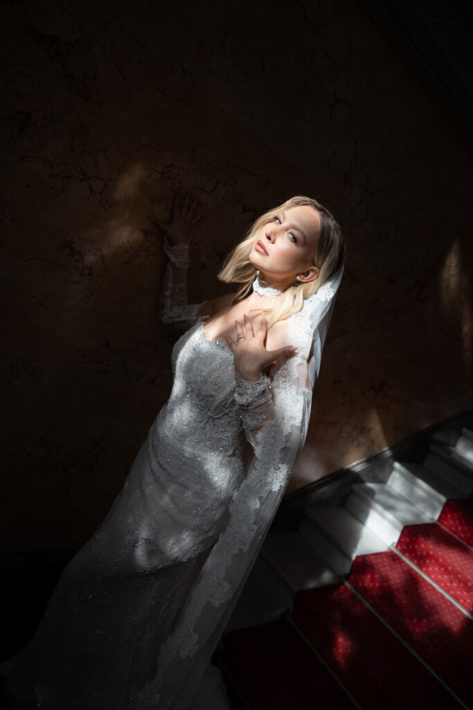 Eine Braut in einem Spitzenkleid und mit Schleier steht auf der Treppe im „Stella Rheni“, blickt nach oben und wird von sanftem Sonnenlicht von oben beleuchtet – ein zauberhafter Moment, perfekt für eine Hochzeit. Heiraten in Bonn war noch nie so bezaubernd.