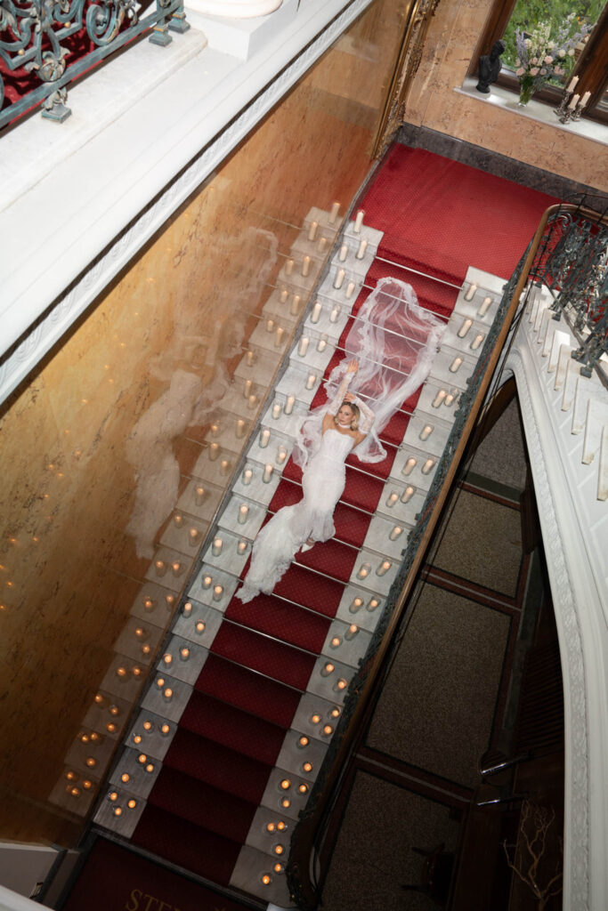 Eine Braut in einem weißen Kleid liegt auf einer mit rotem Teppich ausgelegten, von Kerzen gesäumten Treppe, von oben betrachtet – ein bezaubernder Moment bei Stella Rheni, perfekt für alle, die von einer Hochzeit in Bonn träumen.