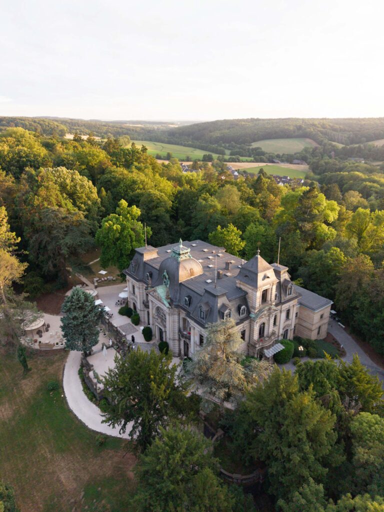 Luftaufnahme von Stella Rheni, einem großen historischen Herrenhaus, umgeben von Bäumen und ländlicher Idylle unter strahlend blauem Himmel – der perfekte Ort für Ihre Hochzeit in Bonn oder um Ihr Heiraten in Bonn in einer bezaubernden Kulisse zu feiern.