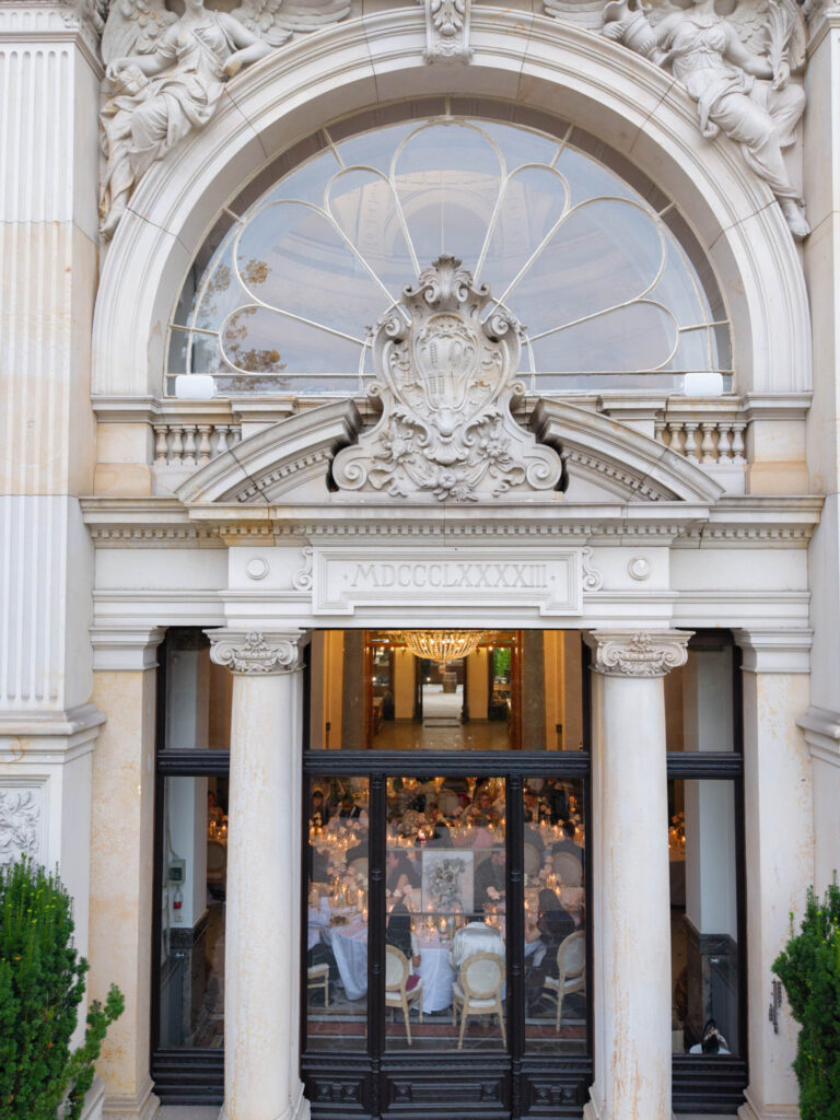 Ein elegantes Gebäude mit kunstvoll verzierten Säulen und einem Bogen, durch dessen große Glastüren man die Gäste beim Essen im Inneren sehen kann – ein perfekter Ort zum Heiraten in Bonn, der jede Hochzeitsfeier in Bonn zu einem wahrhaft unvergesslichen Erlebnis macht.