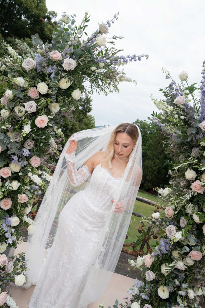 Eine Braut in einem weißen Spitzenkleid steht unter einem Blumenbogen und hält ihren Schleier in der Hand – eine perfekte Inspiration für eine Hochzeit bei Stella Rheni oder für Paare, die darüber nachdenken, in Bonn zu heiraten.