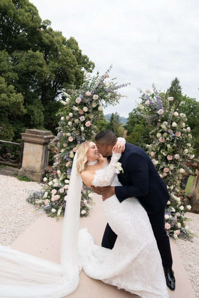 Ein Brautpaar küsst sich vor einem Blumenbogen bei einer Hochzeitszeremonie im Freien und feiert seine Liebe im malerischen Stella Rheni – dem perfekten Ort für Paare, die in Bonn heiraten möchten.