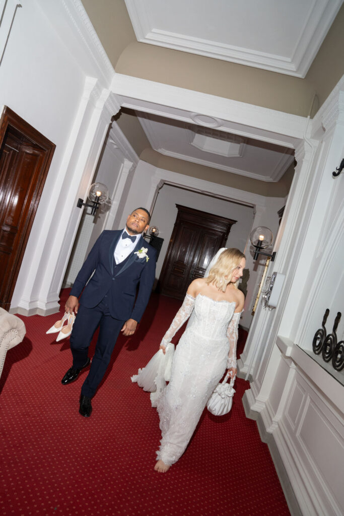 Ein Brautpaar schreitet durch einen Flur im „Stella Rheni“; sie hält ihre Schuhe in der Hand, beide tragen elegante Hochzeitskleidung auf einem roten Teppich – ein Bild, das den Zauber ihrer Hochzeit und die Freude am Heiraten in Bonn einfängt.