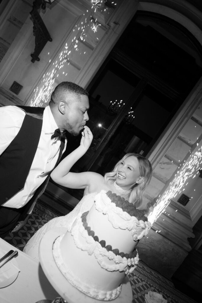 Eine Braut füttert den Bräutigam lachend mit Torte, im Vordergrund sind Wunderkerzen und eine mehrstöckige Torte zu sehen – ein fröhlicher Moment bei ihrer Hochzeit, bei dem sie ihre Liebe feiern, während sie in Bonn im eleganten „Stella Rheni“ heiraten.