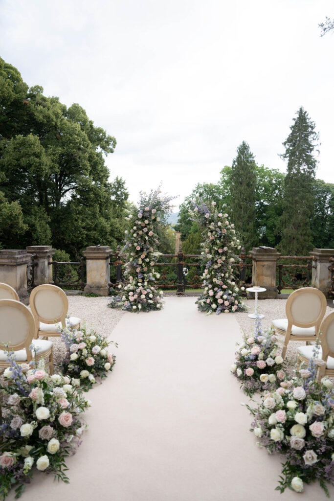 Ein Hochzeitsgang im Freien mit Blumendekorationen, Stühlen mit gerundeter Rückenlehne und üppigem Grün im Hintergrund – perfekt für eine romantische Hochzeit im Stella Rheni. Erleben Sie die Eleganz einer Hochzeit in Bonn inmitten dieser bezaubernden Kulisse.