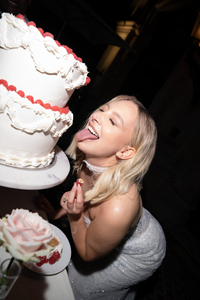 Eine Frau in einem weißen Kleid streckt spielerisch die Zunge heraus – neben einer mit Himbeeren verzierten weißen Torte – und fängt so die unbeschwerte Stimmung einer Hochzeit bei Stella Rheni ein. Wenn Sie planen, in Bonn zu heiraten, spiegelt dieser fröhliche Moment perfekt den Spaß und die Romantik solcher besonderen Feierlichkeiten wider.