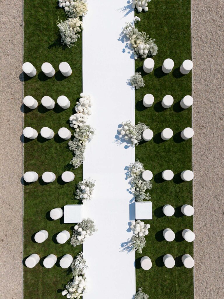 Aerial view of a wedding party with white seating arrangements and floral decorations on green grass, with elegant accents inspired by our wedding collections.