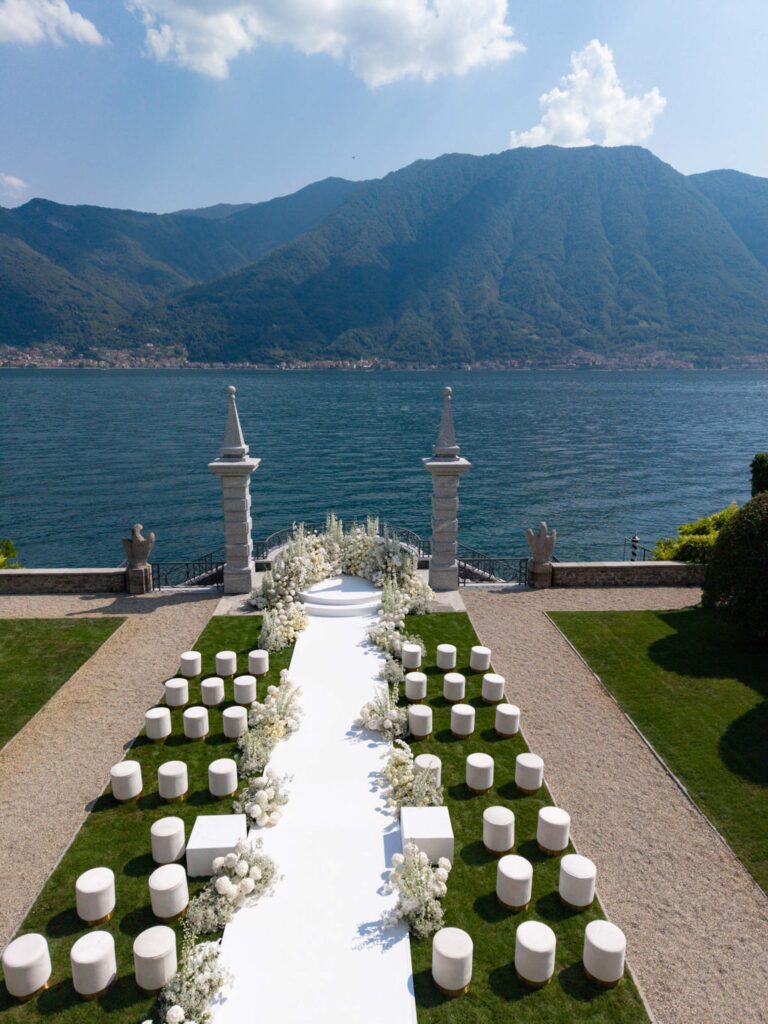A white wedding table with round seating arrangements offers a view of a lake and the mountains under partly cloudy skies, creating a picturesque backdrop inspired by our latest wedding collections.