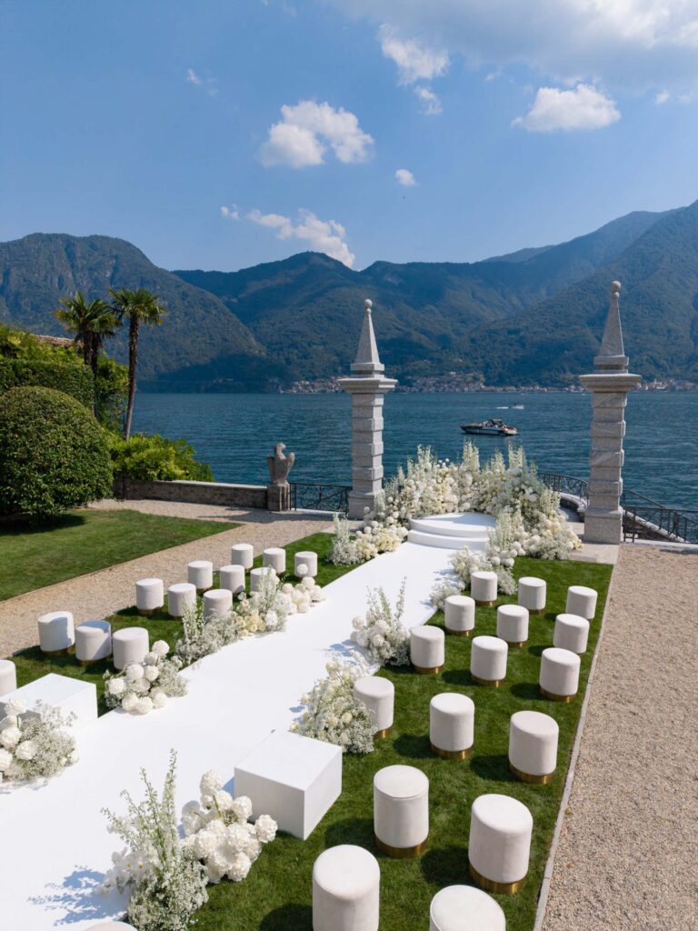 Outdoor wedding decoration with white flowers and wooden benches, overlooking a lake and the mountains under a blue sky, where elements from our wedding collections come to life.