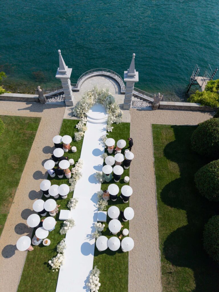 An outdoor wedding decoration by the water with a white aisle, elegant flower arrangements and sunshades for the guests – an exquisite setting, inspired by our wedding collections.