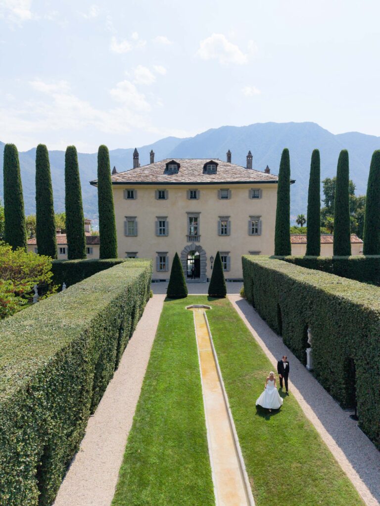 A couple is strolling through a garden with tall hedges, in front of a large villa and with the mountains in the background – a picture that captures the timeless romance that is perfect for wedding collections.