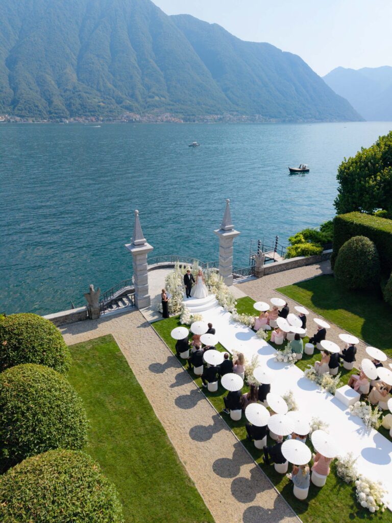 An outdoor wedding at a lake, where the guests sit under white umbrellas surrounded by green mountains in the background – a perfect backdrop for presenting our wedding collections.