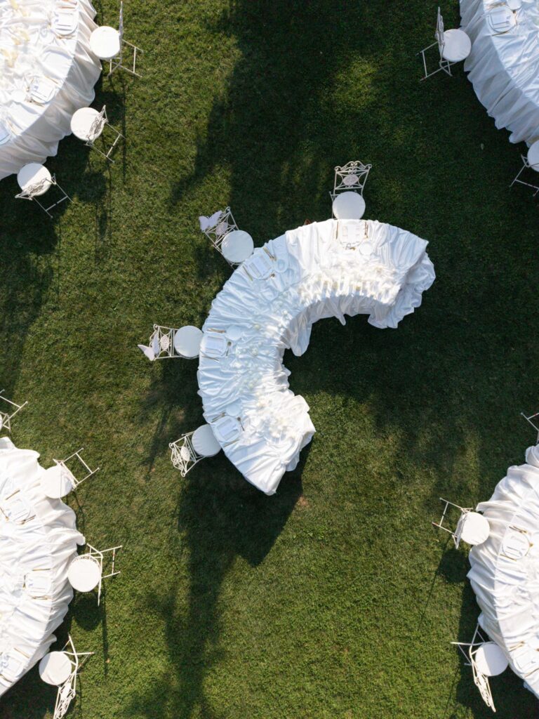 A curved table with a white tablecloth and chairs on a lawn, surrounded by round tables from the „Wedding Collections“ collection, seen from above.