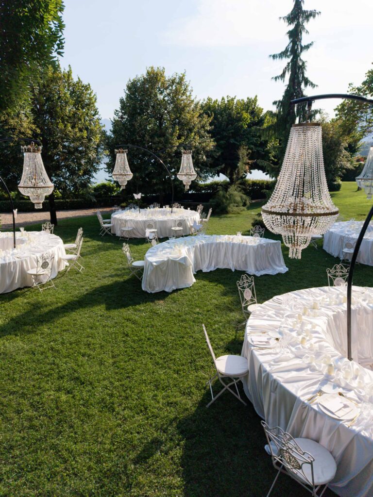 Eine Hochzeitsfeier im Freien mit runden Tischen, die mit weißen Tischdecken gedeckt sind, hängenden Kristallkronleuchtern und einer üppig grünen Rasenfläche – eine elegante Kulisse, inspiriert von unseren Hochzeitskollektionen.