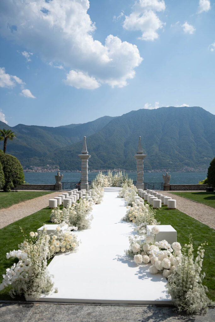 Ein weißer, blumengeschmückter Hochzeitsgang im Freien, mit See und Bergen im Hintergrund unter einem teilweise bewölkten Himmel – das spiegelt die Eleganz unserer Hochzeitskollektionen perfekt wider.