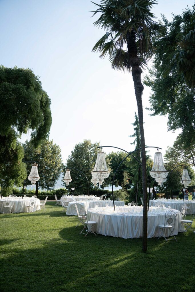 An outdoor event with round tables, white tablecloths, chairs and chandeliers that hang from arches above the lawn – with elegant accents inspired by our wedding collections.