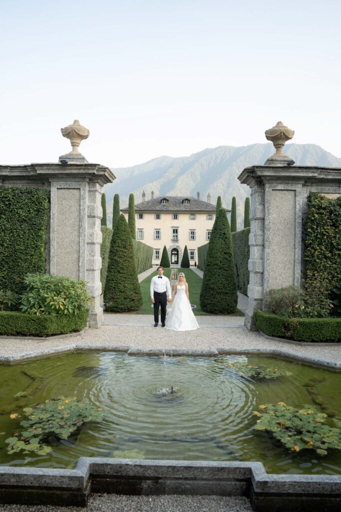 The bride and groom stand in front of a villa, surrounded by manicured hedges, stone columns and a fountain – a breathtaking backdrop that is perfect for capturing unforgettable moments for your wedding photos.
