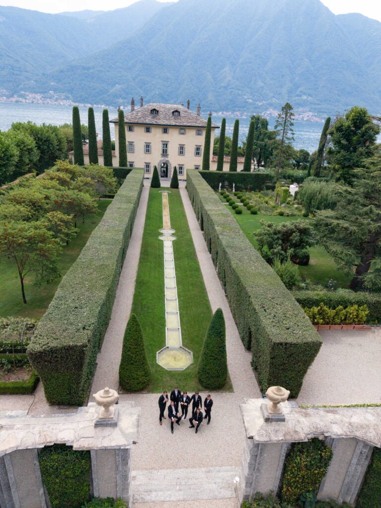 A group of people in festive attire, including stunning wedding dresses from the latest bridal fashion collections, stands in a garden with manicured hedges and a villa overlooking a lake.