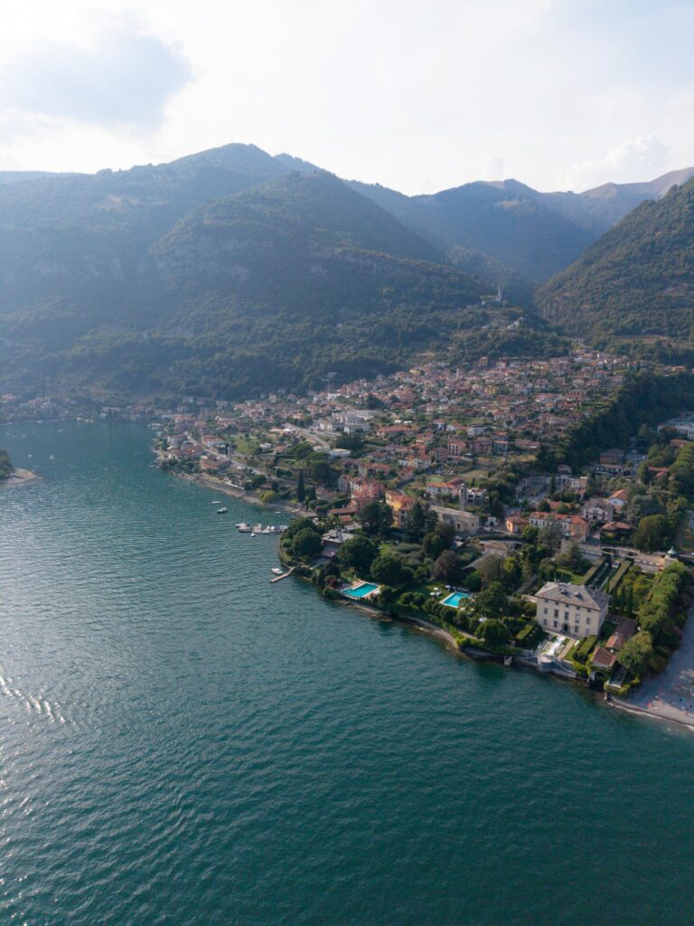 Luftaufnahme einer Stadt am Comer See mit Bergen im Hintergrund und Booten auf dem Wasser, die die atemberaubende Umgebung der Villa Balbiano zeigt – ein unvergesslicher Ort zum Heiraten.
