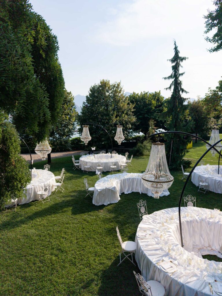 Der Garten der Villa Balbiano mit runden, weiß gedeckten Tischen und Kronleuchtern, umgeben von Bäumen und viel Grün – eine atemberaubende Kulisse im Palazzi-Stil, ideal für eine Hochzeit am Comer See.