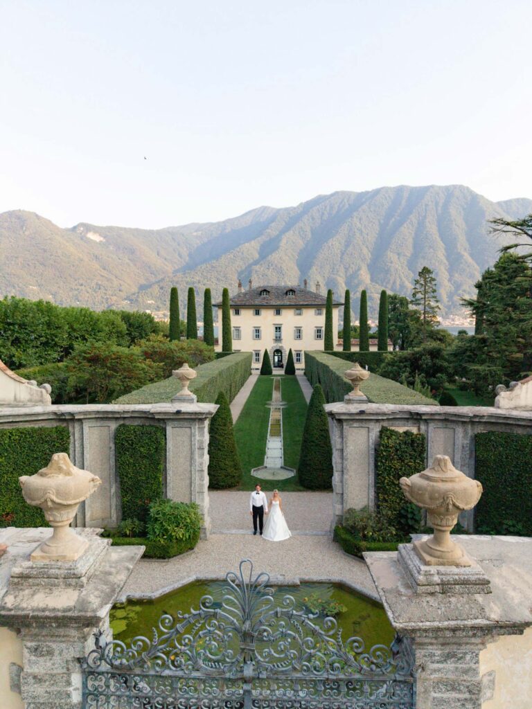 Ein Brautpaar steht in einem gepflegten Garten vor der prächtigen Villa Balbiano, im Hintergrund die majestätischen Berge und das glitzernde Wasser des Comer Sees – eine idyllische Kulisse zum Heiraten.