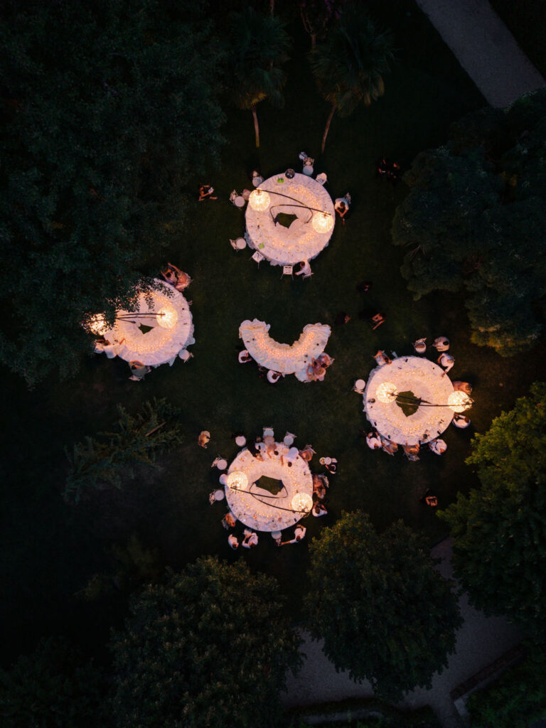 Luftaufnahme von Menschen, die nachts bei warmer Beleuchtung an runden Tischen in einem Garten speisen und einen zauberhaften Abend am Comer See feiern – vielleicht sogar, um in der bezaubernden Villa Balbiano zu heiraten.