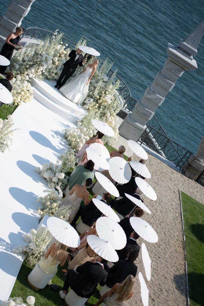Das Brautpaar gibt sich das Ja-Wort bei einer atemberaubenden Hochzeit unter freiem Himmel am Ufer des Comer Sees; die Gäste halten weiße Sonnenschirme entlang des Ganges und unterstreichen so die romantische Eleganz, für die die Villa Balbiano bekannt ist.