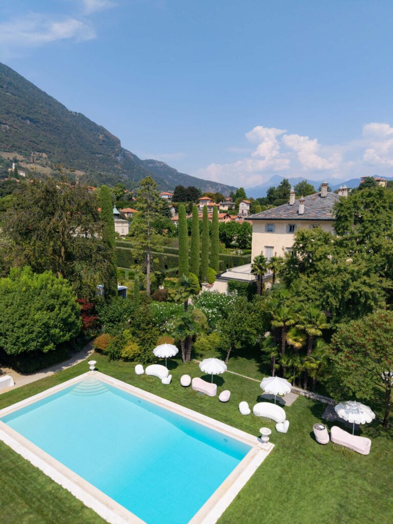 Ein rechteckiger Pool mit weißen Sonnenschirmen und Liegestühlen, umgeben von grünen Gärten und fernen Bergen, versetzt einen in die elegante Atmosphäre der Palazzi am Comer See – ideal für alle, die davon träumen, am Comer See in einer atemberaubenden Kulisse am Seeufer zu heiraten, die an die Villa Balbiano erinnert.