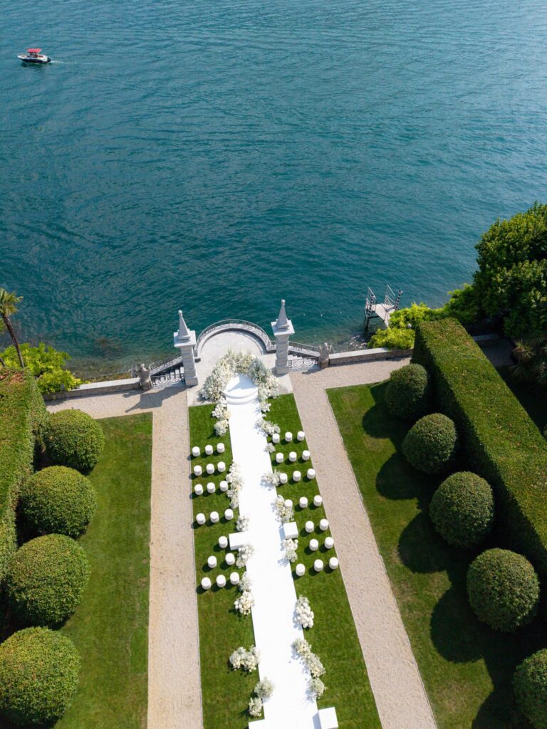 Luftaufnahme eines Hochzeitsgangs mit weißen Stühlen am Ufer der atemberaubenden Villa Balbiano am Comer See, umgeben von grünen Hecken und Rasenflächen – eine unvergessliche Kulisse, um stilvoll zu heiraten.