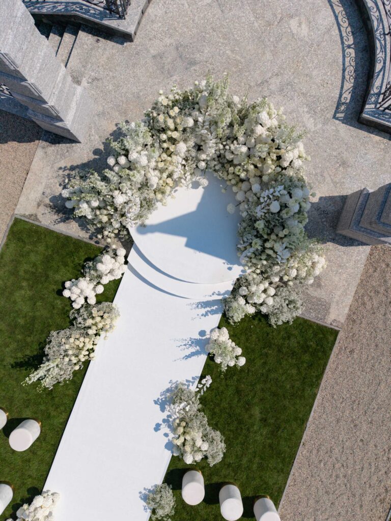 Luftaufnahme eines weißen Hochzeitsgangs mit Blumenbögen und weißen Blumen, umgeben von grünem Gras – eine bezaubernde Kulisse für eine Hochzeit am Comer See, mit der prächtigen Villa Balbiano und historischen Palazzi als atemberaubende Kulisse.