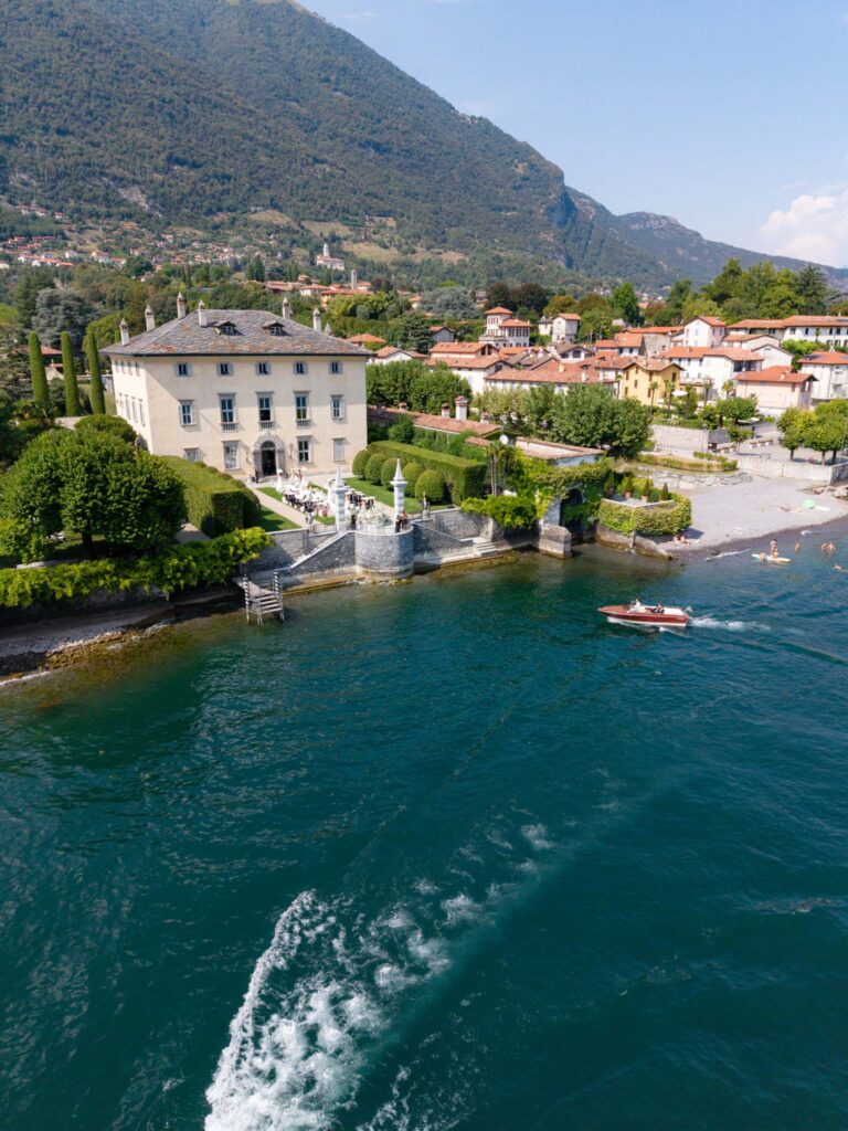 Eine Villa am See, mit Bergen im Hintergrund, Booten auf dem Wasser und einem kleinen Strand mit Menschen in der Nähe – das erinnert an die Pracht von Palazzi wie der Villa Balbiano, eine idyllische Kulisse für alle, die davon träumen, am Comer See zu heiraten.