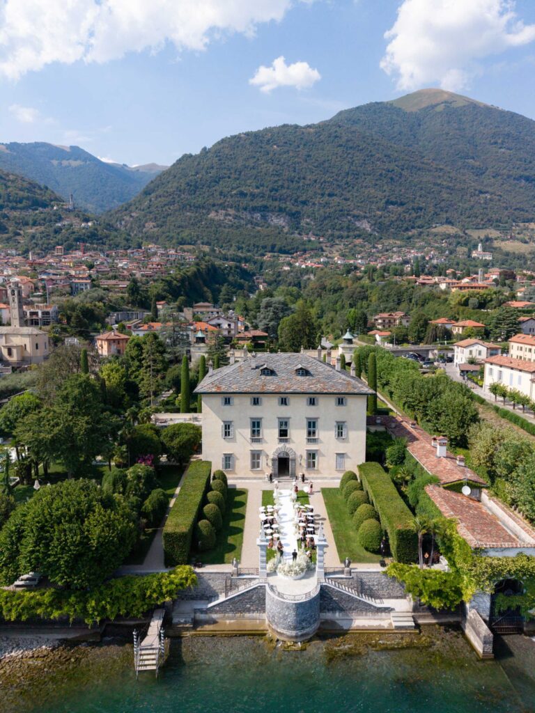 Am Seeufer liegt eine prächtige Villa mit Gärten, die an die berühmten Palazzi am Comer See erinnert, mit Bergen und einer Stadt im Hintergrund unter blauem Himmel – eine wahrhaft malerische Kulisse für alle, die davon träumen, am Comer See zu heiraten.