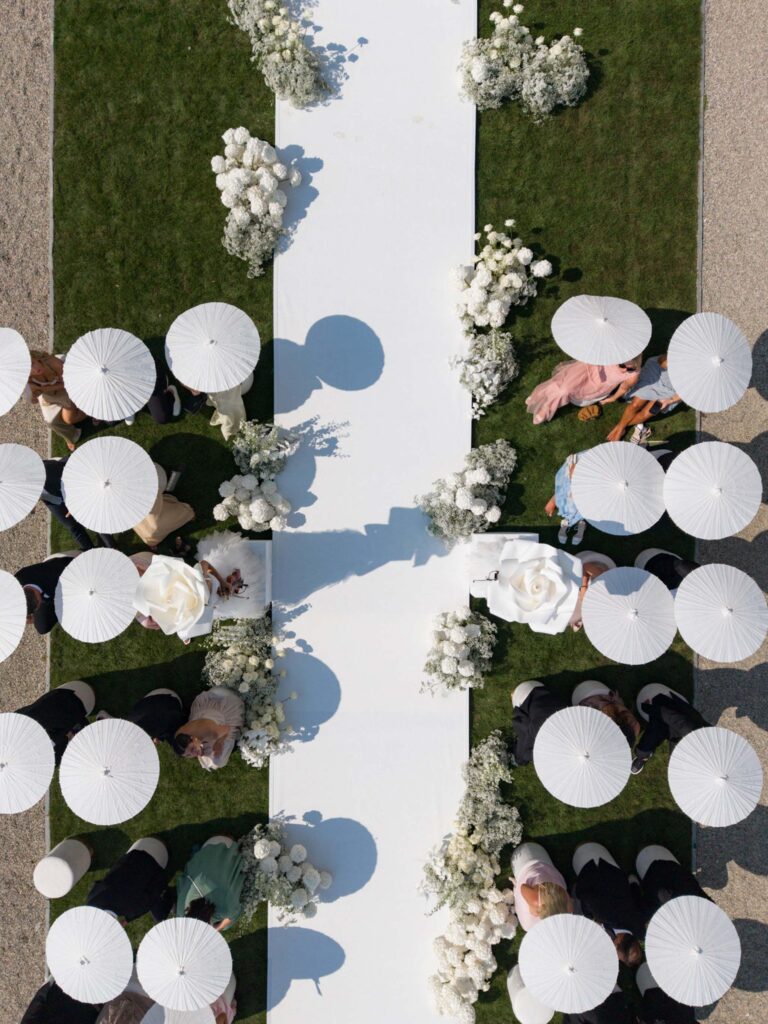 Luftaufnahme eines Hochzeitsgangs in der Villa Balbiano am malerischen Comer See, mit Gästen, die weiße Regenschirme in den Händen halten, und eleganten weißen Blumendekorationen – ein unvergesslicher Ort zum Heiraten.