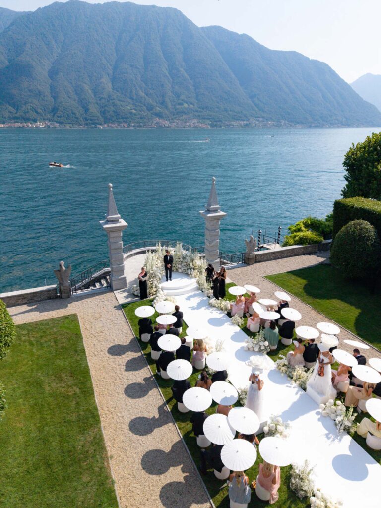 Eine Hochzeitszeremonie im Freien am Seeufer, bei der die Gäste weiße Regenschirme in den Händen halten und der Blick auf Berge und Wasser fällt – eine bezaubernde Kulisse, die an die Villa Balbiano und die zeitlose Eleganz der Palazzi am Comer See erinnert, perfekt für alle, die davon träumen, am Comer See zu heiraten.