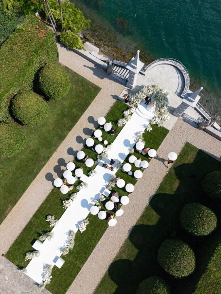 Luftaufnahme einer Hochzeitszeremonie im Freien mit weißen Stühlen und einem Gang am Wasser in der atemberaubenden Villa Balbiano mit Blick auf den ruhigen Comer See – ein perfekter Rahmen, um auf unvergessliche Weise zu heiraten.
