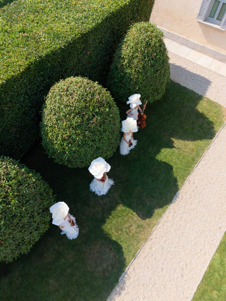 Vier Personen in weißen Kleidern und mit großen weißen Hüten stehen neben gepflegten Sträuchern auf dem Rasen der Villa Balbiano, einem atemberaubenden Veranstaltungsort am Comer See, der sich perfekt für alle eignet, die in einer bezaubernden italienischen Kulisse heiraten möchten.