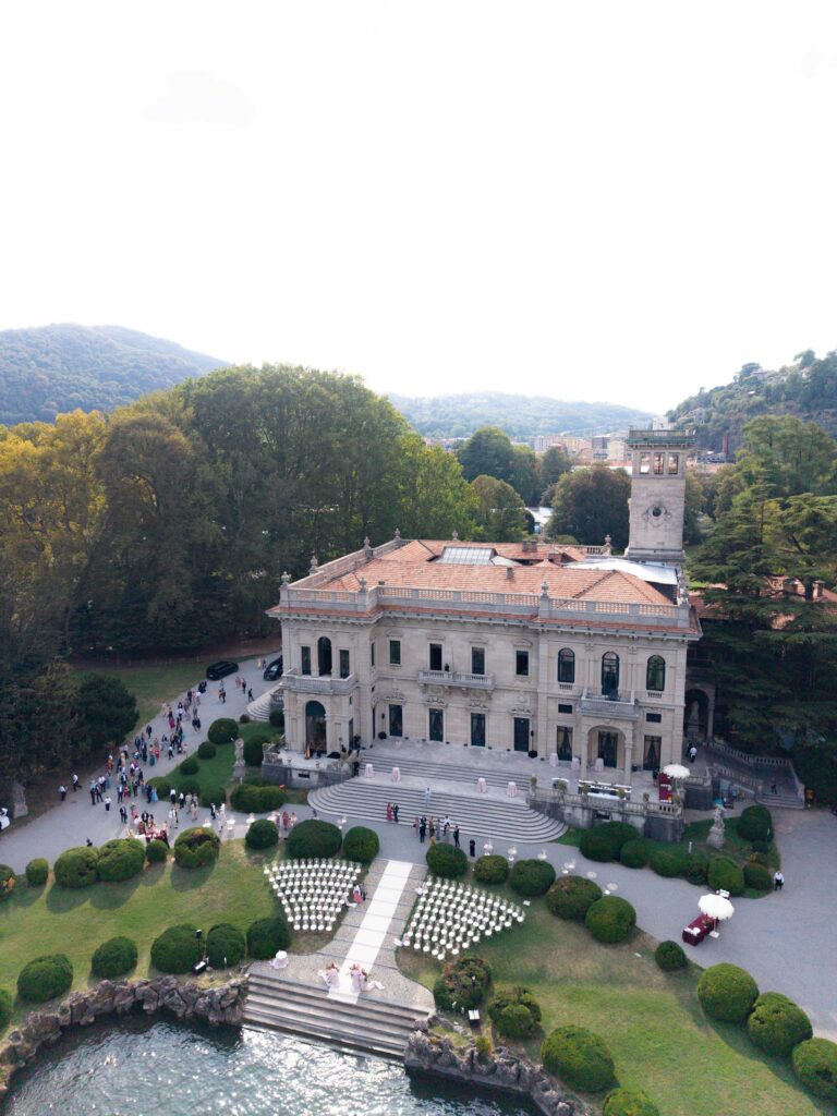 Luftaufnahme einer prächtigen Villa am See mit Gärten, versammelten Menschen und automatischem Entwurf voller Eleganz.