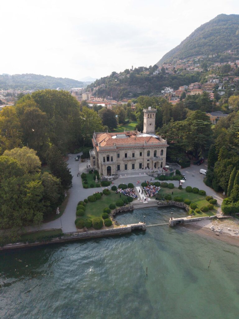 Luftaufnahme einer Villa am See mit Gärten, Bäumen, Hügeln in der Ferne und einer Automatischer Entwurf-Veranstaltung auf dem Rasen.