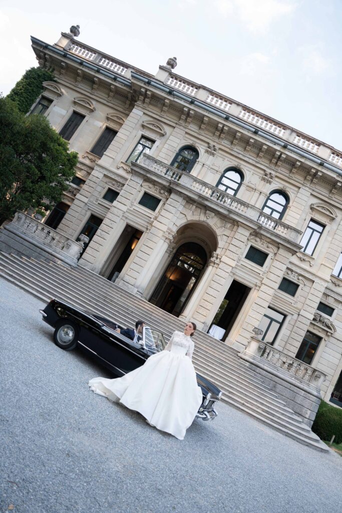 Eine Braut in einem weißen Kleid steht neben einem schwarzen Oldtimer und verkörpert die Eleganz von Automatischer Entwurf.