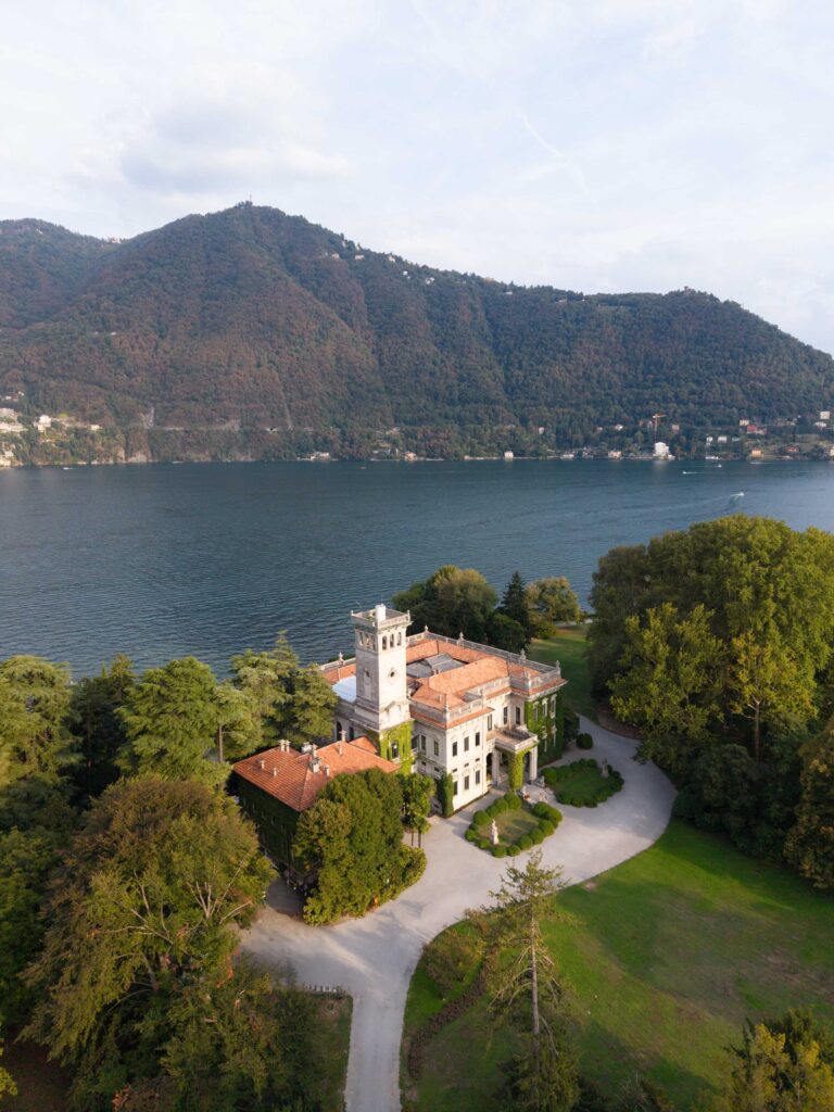 Eine große Villa mit einem Turm am Seeufer, ein automatischer Entwurf, umgeben von Bäumen und Bergen.