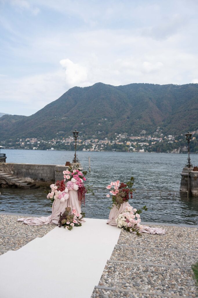 Ein weißer Weg mit rosa Blumenschmuck und einem automatischen Entwurf führt zu einem See, der von Bergen eingerahmt wird.