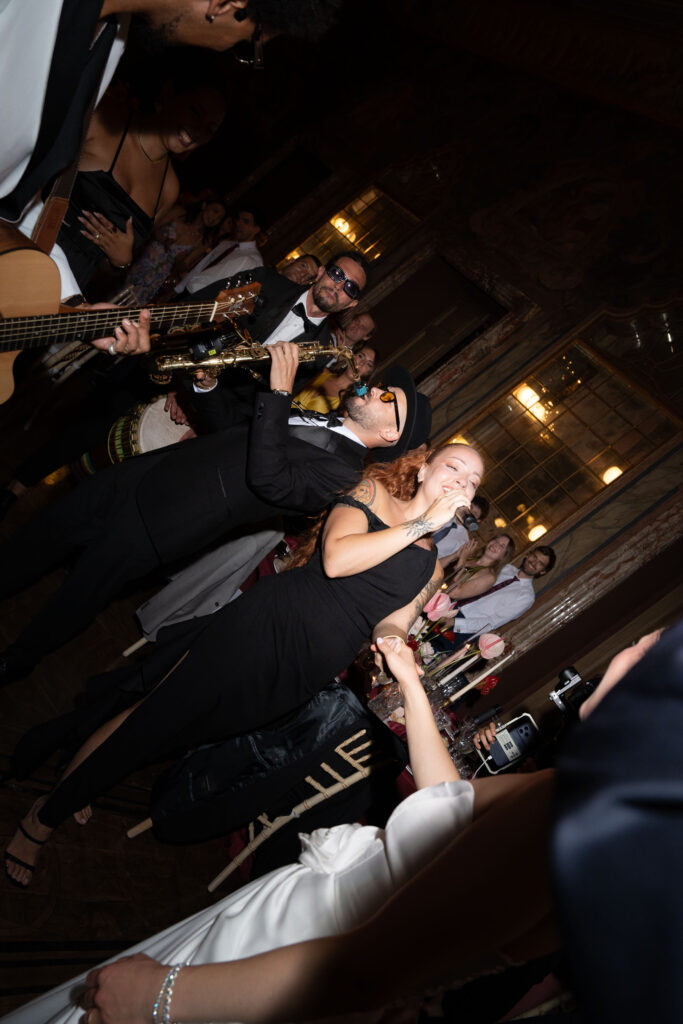 Eine Frau singt und lächelt zusammen mit einer Band bei einer Veranstaltung in einem Saal, umgeben von einer Atmosphäre voller Eleganz.